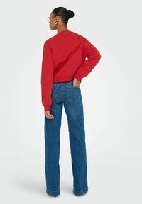 Vrouw met een bun-haarstijl, die een bril draagt, een rode trui en een blauwe wijde broek aan heeft, staande met één hand in haar achterzak.
