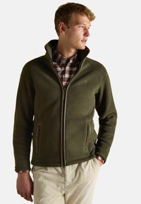 Olivovo zelená fleecová bunda s vysok�ým golierom, zipsom vpredu a vreckami. Nosená cez kockovanú košeľu, s rebrovanými manžetami na rukávoch.