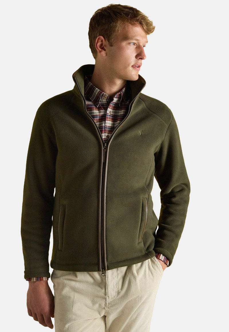 Olivovo zelená fleecová bunda s vysokým golierom, zipsom vpredu a vreckami. Nosená cez kockovanú košeľu, s rebrovanými manžetami na rukávoch.