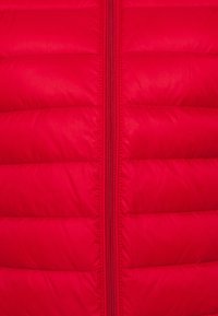 Veste matelassée rouge avec texture matelassée, dotée d'une fermeture éclair centrale et d'un tissu lisse et brillant. Le design est épuré avec des panneaux carrés.