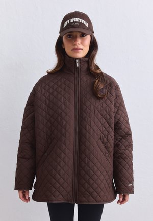 Donna che indossa una giacca trapuntata marrone con zip e un cappellino da baseball marrone coordinato con testo bianco, in piedi davanti a una parete chiara semplice.