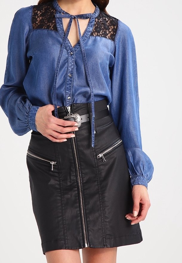 Femme portant un chemisier bleu orné de dentelle avec un col noué et une jupe en cuir noire taille haute avec des fermetures éclair et une boucle de ceinture argentée.