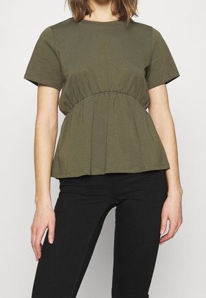 Frau trägt ein olivgrünes Kurzarm-Top mit geraffter Taille, kombiniert mit enganliegenden schwarzen Hosen vor einem schlichten Hintergrund.