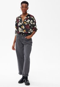 Femme se tenant debout, les mains dans les poches, portant une chemise à boutons noire à fleurs, un pantalon taille haute gris, une ceinture noire et des mocassins noirs avec des chaussettes.