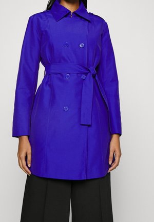 Femme portant un trench-coat bleu vif à double boutonnage avec ceinture à la taille et un pantalon noir à jambes larges, debout devant un fond uni.