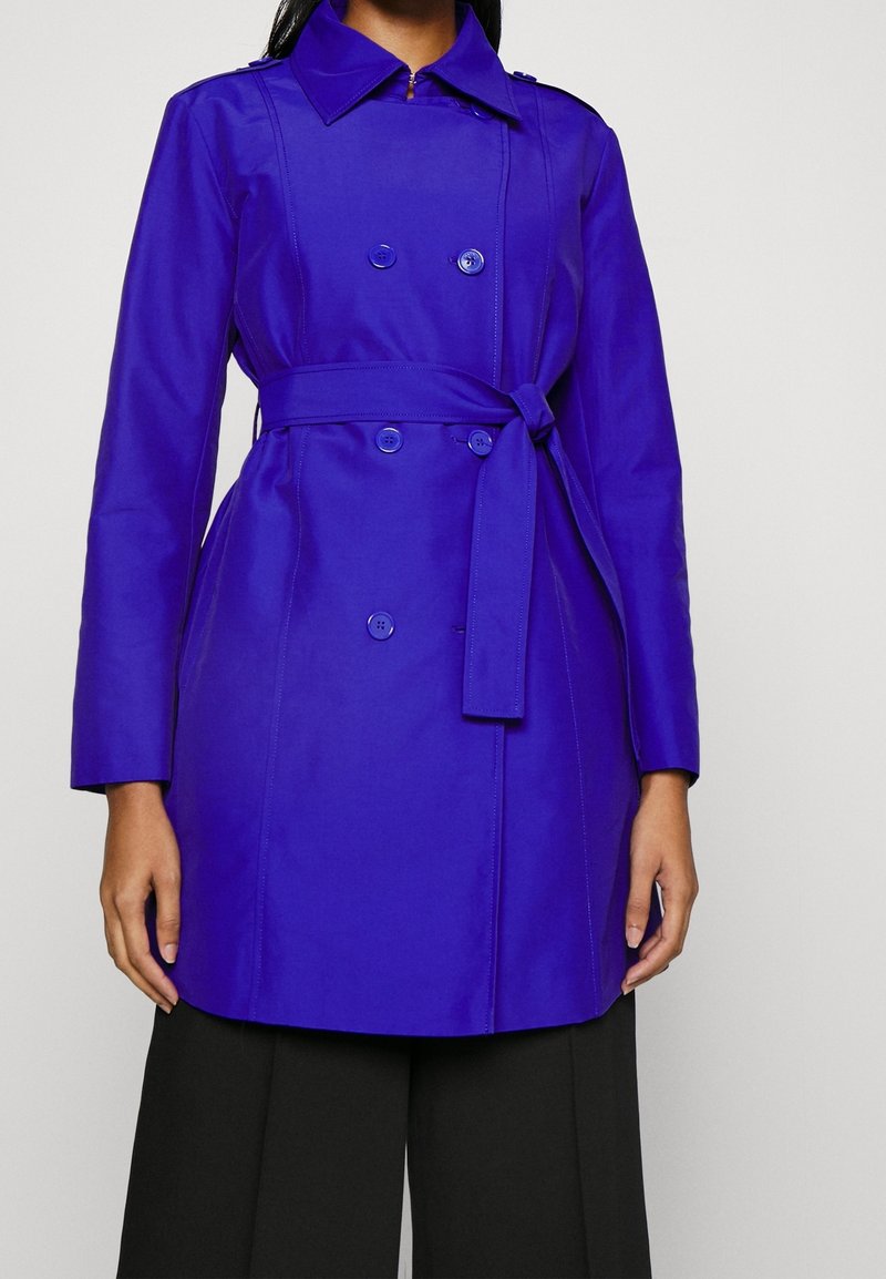 Femme portant un trench-coat bleu vif à double boutonnage avec ceinture à la taille et un pantalon noir à jambes larges, debout devant un fond uni.