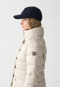 Chaqueta de plumas beige con cuello alto y textura acolchada. Presenta un logo circular en el brazo izquierdo. Gorro azul oscuro con visera.