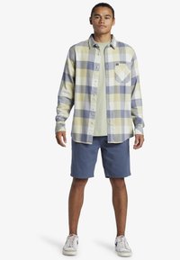 Gekleurde flanellen overhemd in geel, grijs en blauw met een knoopsluiting, voorzien van een linker borstzak. Gecombineerd met blauwe shorts en sneakers.