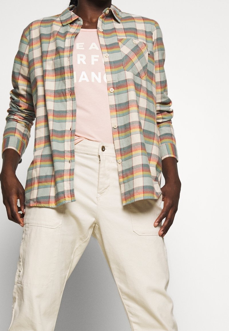 Flannel shirt featuring blue, green, and orange stripes, with a breast pocket. Worn over a pink t-shirt and paired with light beige pants.