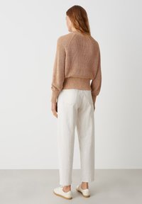 Hellbrauner Strickpullover mit geripptem Saum und puffärmel, kombiniert mit weißen, locker sitzenden Hosen und hellen Schuhen.