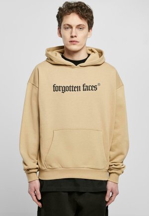 Beige hettegenser med lommer foran og snor i hetta. Svart tekst "glemte ansikter" trykt på brystet. Mykt stoff med en avslappet passform.
