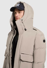 Light beige puffer jacket with a large hood, featuring black accents and multiple pockets; made from smooth, insulated material.