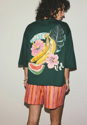 Osoba o kręconych włosach ubrana w ciemnozieloną koszulę z grafiką bananów, różowych kwiatów i arbuza, zestawioną z pomarańczowymi szortami w paski.