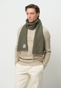 Foulard verde oliva realizzato in morbido tessuto, con dettagli frangiati alle estremità e un piccolo tag con il logo. Indossato sopra un maglione beige.