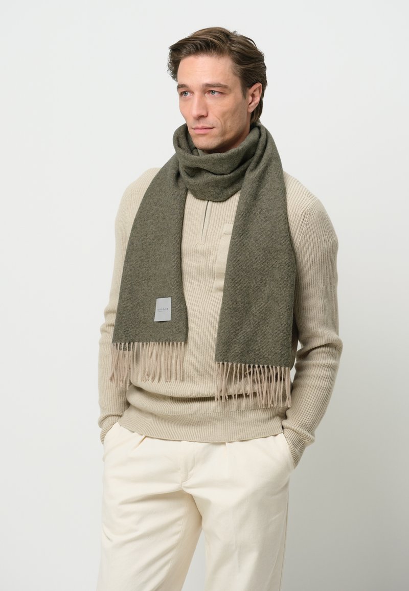 Bufanda verde oliva hecha de tela suave, con detalles de flecos en los extremos y una pequeña etiqueta con el logo. Llevada sobre un suéter beige.