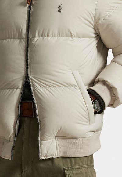 Casaco puffer bege com fecho à frente, punhos canelados e bolsos laterais. Apresenta um pequeno logótipos e um acabamento texturizado.