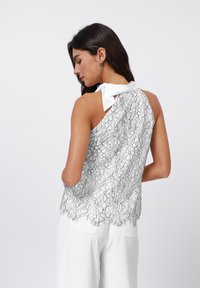 White top with a floral lace pattern, featuring a back tie detail. The hem is scalloped, and it has a slightly sheer texture.