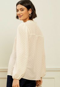 Blusa leggera e trasparente con motivo a pois, maniche a sbuffo e colletto arricciato. Colore crema con una silhouette fluida, abbinata a pantaloni scuri.