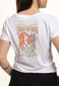 T-shirt en coton blanc présentant une illustration colorée d'une sirène aux cheveux rouges, ornée de fleurs et de accents dorés, étiquetée "Fille de Triton".