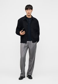 Mann mit klaren Brillengläsern, schwarzer Jacke, dunkelblauem Polohemd, grauer Hose und schwarzen Schuhen, der vor einem schlichten Hintergrund eine schwarze Geldbörse hält.