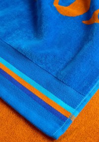 Asciugamano blu con design arancione, caratterizzato da una superficie testurizzata. I bordi presentano strisce blu, arancioni e turchesi. Tessuto morbido, forma rettangolare.