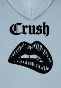 Gaiši zila kapuce ar lielu melnu izšūtu tekstu "Crush" virs ilustrēta atvērta mutes dizaina ar detaļām par lūpām un zobiem.