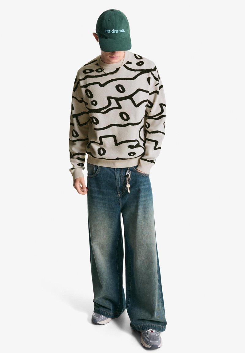 Pulover v bež pletenini z črnim abstraktnim vzorcem, v kombinaciji s širokimi jeans hlačami in sivimi supergami. Zelen klobuk z belim napisom.