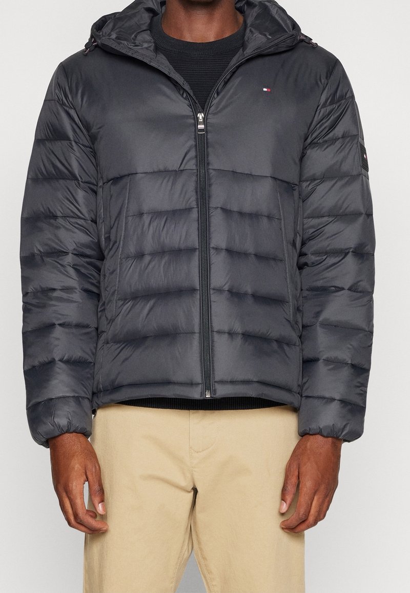 Homme portant une veste matelassée noire à capuche avec fermeture éclair devant, par-dessus une chemise noire et un pantalon beige, debout devant un fond clair.