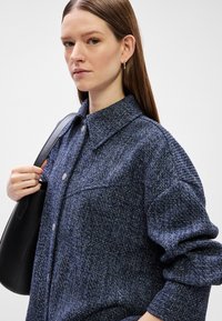 Camisa oversized texturizada azul com colarinho e fecho de botão. Saco de ombro preto segurado com uma mão.