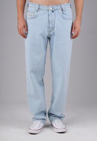 Person, die hellblaue Jeans und weiße Sneakers trägt und vor einem schlichten grauen Hintergrund steht.