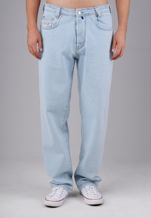 Person, die hellblaue Jeans und weiße Sneakers trägt und vor einem schlichten grauen Hintergrund steht.