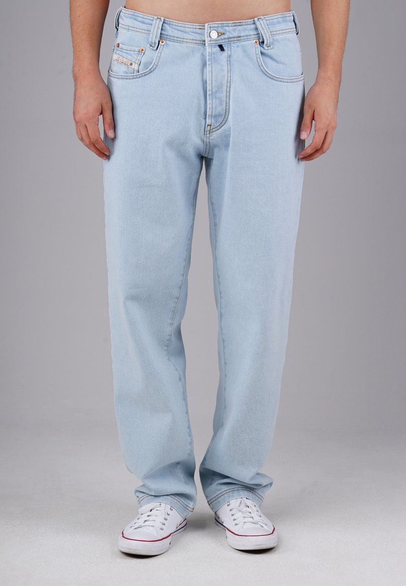 Person, die hellblaue Jeans und weiße Sneakers trägt und vor einem schlichten grauen Hintergrund steht.