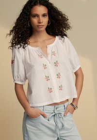 Hvid bluse med puffede ærmer, prikket tekstur, blomsterbroderi i pink og grøn, knaplukning foran, cropped design. Båret med lyseblå shorts.