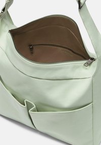 Bolsa de tela suave de color verde claro con cierre de cremallera y bolsillos interiores. Presenta dos bolsillos exteriores y una textura lisa. El interior es marrón.