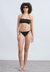 Μαύρο μπικίνι bandeau με adjustable tie-side bottoms. Λείο ύφασμα, ελάχιστη κάλυψη, με λεπτές τιράντες και απλό σχέδιο.