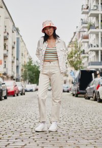 Cream denim jacket over a striped tank top, cream trousers, and white sneakers. A light pink bucket hat with orange accents. Cobblestone street.