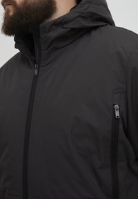 Jachetă neagră impermeabilă cu glugă, prevăzută cu fermoar frontal și buzunar lateral cu fermoar. Textură netedă și design minimalist.