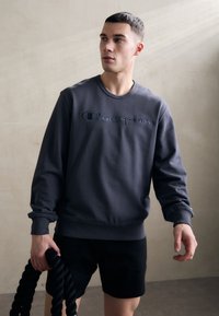 Grauer Rundhals-Sweatshirt aus weichem Material mit gesticktem "Champion"-Logo. Kombiniert mit schwarzen Shorts und hält ein schwarzes Seil.