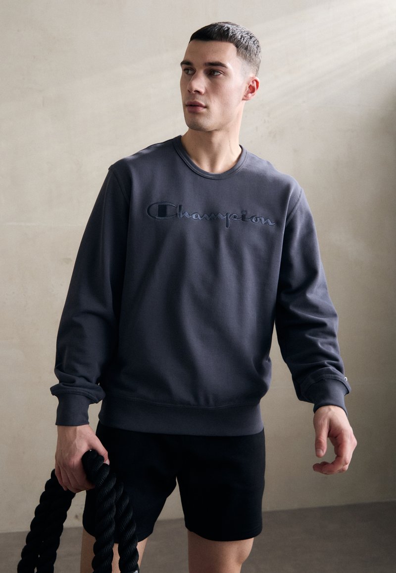 Grauer Rundhals-Sweatshirt aus weichem Material mit gesticktem "Champion"-Logo. Kombiniert mit schwarzen Shorts und hält ein schwarzes Seil.