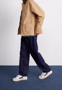 Ljusbrun jacka med brun krage, framfickor, bärs över en blå skjorta. Mörkblå byxor och sneakers med vita, svarta och beige accenter.