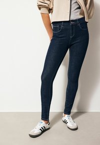 Jeans skinny de denim azul oscuro con costuras claras, dos bolsillos delanteros, cintura alta y combinados con zapatillas deportivas en blanco y gris.