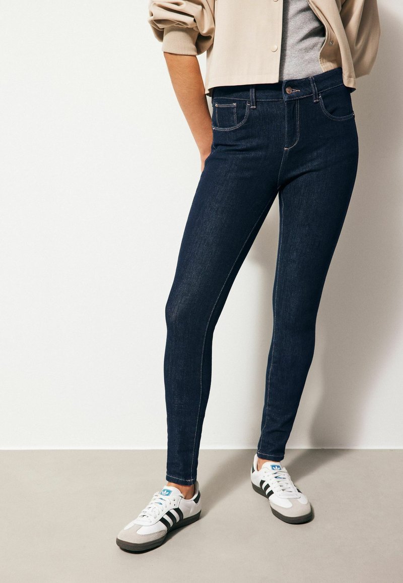 Jeans skinny de denim azul oscuro con costuras claras, dos bolsillos delanteros, cintura alta y combinados con zapatillas deportivas en blanco y gris.