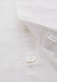 Witte button-up shirt met een gladde textuur, voorzien van een ronde kraag en een enkele witte knop bij de hals.