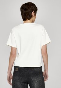 Krótki rękaw, t-shirt z okrągłym dekoltem w miękkim białym materiale, o prostym wzornictwie. W połączeniu z czarnymi spodniami z perforowanymi detalami i logo.