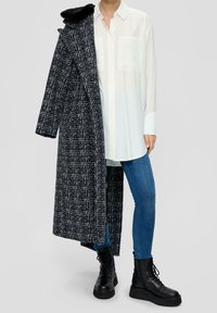 Long, black and white check pattern coat with fur collar, layered over white button-up shirt. Blue jeans and black lace-up combat boots complete the look.
