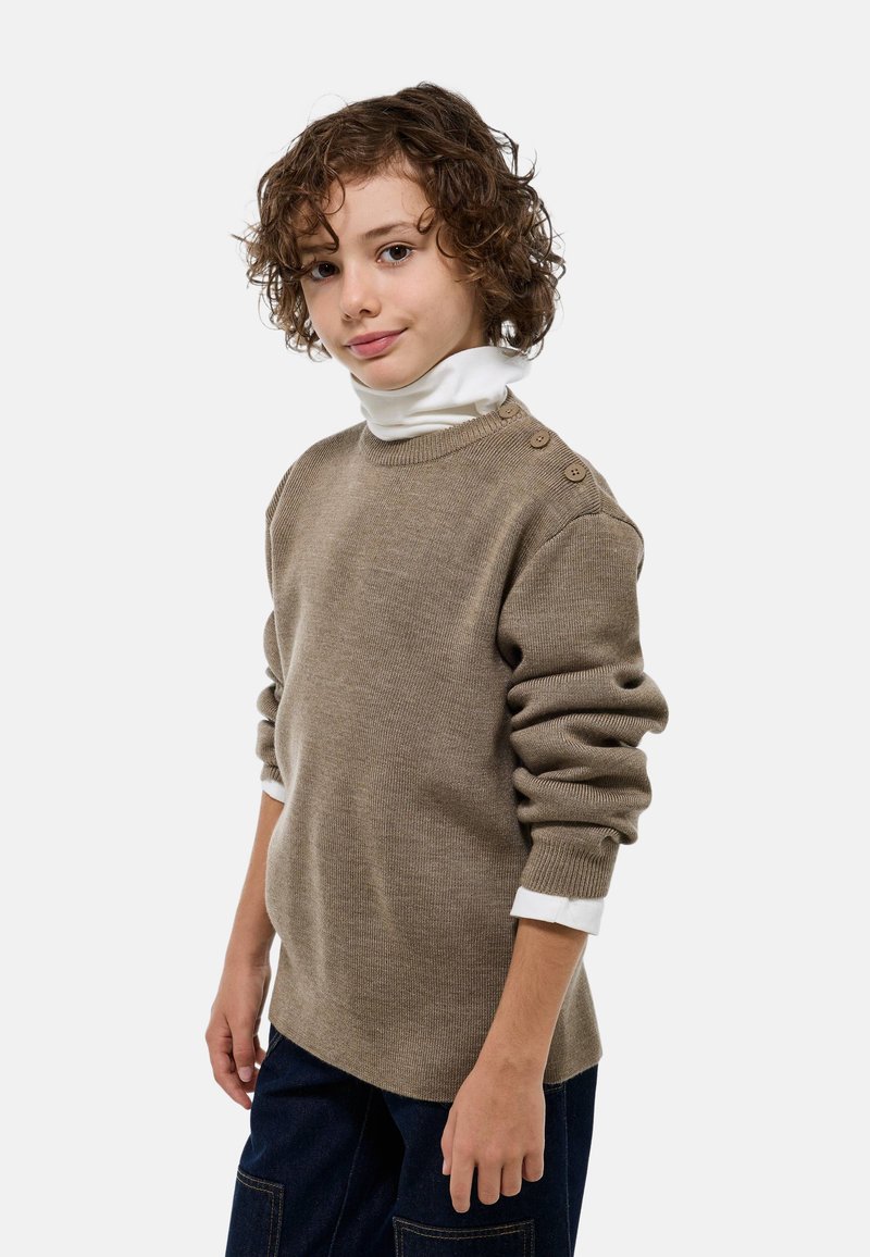 Pull en tricot brun avec un décolleté large, tissu texturé et trois boutons sur l'épaule gauche, superposé à un col roulé blanc.