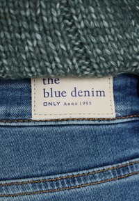 Blue denim jeans with a woven green sweater above. A label reads “the blue denim ONLY Anno 1995” in dark blue on a cream background.