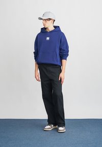 Uma hoodie azul com uma gráfica na parte da frente, calças pretas e um boné cinza. A hoodie tem um bolso canguru e punhos canelados, enquanto os sapatos apresentam detalhes claros.