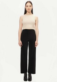 Gräddfärgad ribbad linne tillsammans med högmidjade svarta kordflamsbyxor med vida ben. Modellen bär svarta ankelboots och poserar mot en vit bakgrund.