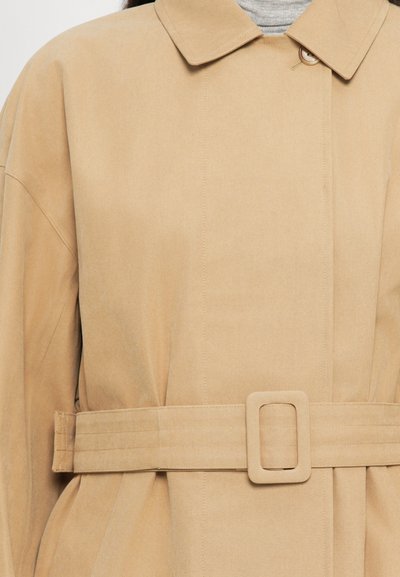 Közelkép egy bézs színű trench kabátról, széles övvel, amely téglalap alakú csattal ellátott, szürke garbó pulóver fölött viselve.